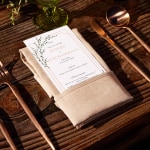 Un menu de mariage moderne repose sur une serviette beige sur une table en bois.