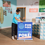 Au cœur d’un restaurant animé, un homme installe une banderole rétractable pour présenter ses bols de poké.