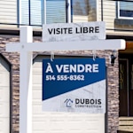 Gros plan sur un ajout annonçant une visite libre, fixé en haut d’un poteau potence blanc, devant une maison.