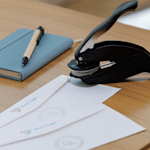 Un bloc-notes, un stylo et une pince à gaufrer sont posés sur un bureau, à côté de deux enveloppes blanches, chacune marquée d’une impression ronde gaufrée.