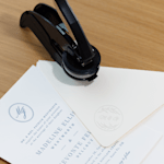 Een reliëfstempel en bruiloftsdrukwerk naast elkaar geplaatst, waar met de stempel een bijpassende reliëfafdruk op de gepersonaliseerde envelop is gemaakt.