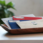 Diferentes envelopes coloridos empilhados num tabuleiro com correspondência sobre uma mesa de madeira.