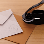 Um carimbo de alto relevo ao lado de envelopes em papel kraft com um selo branco com monograma, dispostos sobre uma superfície de madeira.