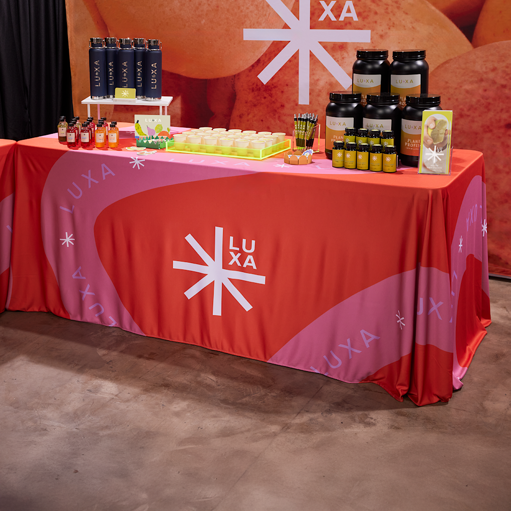 A display table with Luxa branded products and supplements, covered by an orange and pink tablecloth at a trade show.