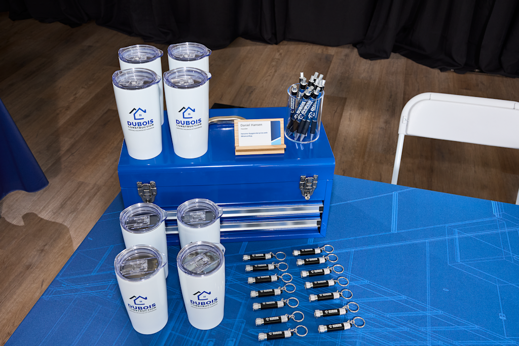 A trade show display table with Dubois Construction branded tumblers, business cards, roller banners, and promotional keychains on blue tablecloth.

